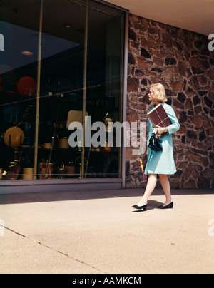 1960 anni sessanta DONNA BIONDA Blue Coat passeggiando per le strade dello shopping della finestra CERCA IN STORE NEGOZIO DI VENDITA AL DETTAGLIO DI WINDOWS retrò Foto Stock
