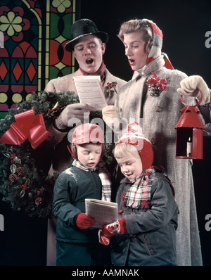 Anni Cinquanta famiglia 4 cantando canti natalizi papà UOMO HA CORONA SU BRACCIO DONNA MOM trattiene lanterna rossa ragazza ragazzo Musica La canzone NEVE OUTDOOR Foto Stock