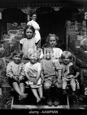 1890s 1900 svolta del secolo GRUPPO DI SEI sporco bambini seduti sulle scale davanti a casa con la madre in piedi sul portico Foto Stock