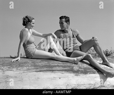 1930s coppia giovane IN COSTUME DA SEDUTI vicino alla spiaggia sulla barca capovolta Foto Stock
