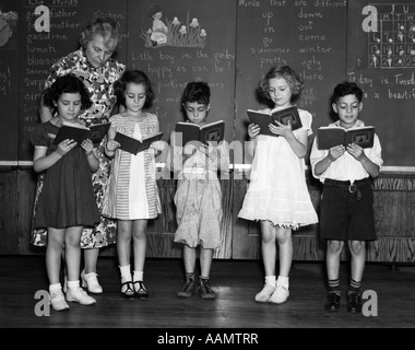 1930s line-up di 5 GLI STUDENTI DELLA SCUOLA ELEMENTARE DI FRONTE BLACKBOARD la lettura di libri con insegnante cercando su Foto Stock