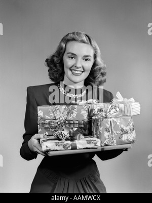 Anni Cinquanta donna sorridente HOLDING pila di avvolgere i regali di Natale guardando la fotocamera Foto Stock