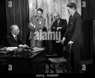 1920s 1930 RIUNIONE DEL perplesso di uomini in abiti riuniti intorno a scrivania in ufficio business Foto Stock