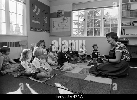 Anni ottanta GRADE SCHOOL insegnante seduto sul pavimento con gli studenti a suonare la chitarra e cantare Foto Stock