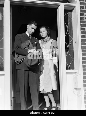 1930s tra marito e moglie di stand in open door man andando al lavoro controllo guardare la valigetta di contenimento cappello in mano Foto Stock