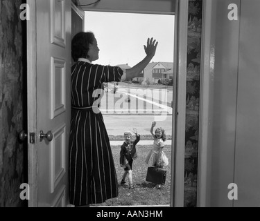 Anni Cinquanta donna madre sventolando dalla porta come figlio del ragazzo e ragazza figlia lasciare PER LA SCUOLA DI MATTINA Foto Stock