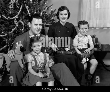 Negli anni quaranta anni cinquanta famiglia seduti intorno ad albero di Natale azienda doni guardando la fotocamera Foto Stock