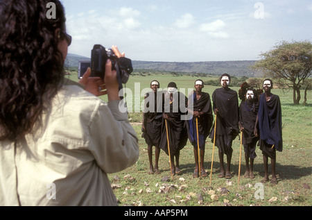 Sei giovani uomini Maasai con mantelli di colore nero e bianco di fronte vernice essendo girato da un turista bianco Tanzania Foto Stock