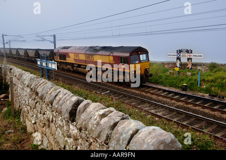 Un EWS freight attraversa il confine dalla Scozia in Inghilterra su un treno di carbone passando un vecchio British Railways segno con bandiere Foto Stock
