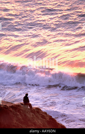 Uomo seduto sulla spiaggia rocciosa guardando le onde dell'oceano crash sulla spiaggia al tramonto Foto Stock