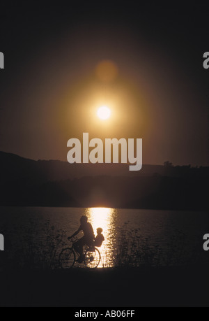 Silhouette di una bicicletta padre con il bambino nel seggiolino per bambini passando da un lago che riflette il sole di setting Foto Stock