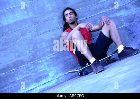 Giovane uomo seduto su skateboard contro un muro di cemento Foto Stock