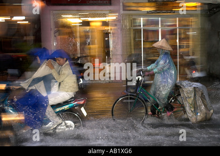 La donna nel cappello conico e cape corse in bicicletta lungo allagato De Tham Street Pham Ngu Lao district Ho Chi Minh City Vietnam Foto Stock