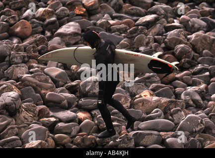 Un surfista in una muta attraversando una spiaggia rocciosa vicino a North Devon VILLAGGIO DI PESCATORI DI CLOVELLY REGNO UNITO Foto Stock