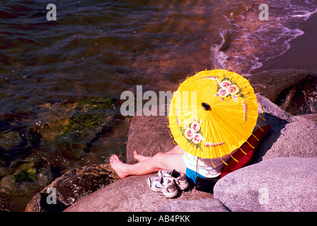 Persona seduta su una roccia sulla spiaggia rocciosa lungo il bordo delle acque e prendere il sole sotto Carta gialla Ombrellone / Parasol Foto Stock