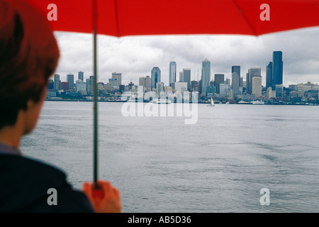 Donna con ombrello rosso a piedi sotto la pioggia lungo la riva del Puget Sound con Seattle Washington Skyline a distanza Foto Stock
