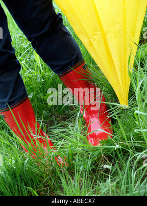 Signor gambe di una persona della donna con red rubberboots sotto un ombrello giallo di andare a fare una passeggiata nel tempo piovoso su un verde prato umido Foto Stock