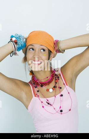 Ragazza adolescente indossando un sacco di gioielli, mani dietro la testa, guardando in alto, sorridente, ritratto Foto Stock