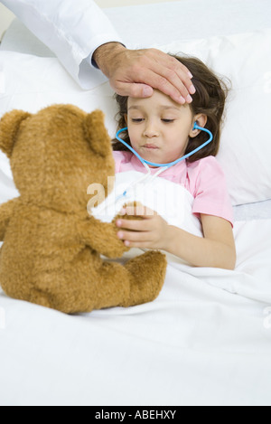 Ragazza distesa nel letto di ospedale, tenendo Teddy bear, medico la mano sulla fronte Foto Stock