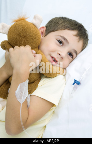 Ragazzo che giace nel letto di ospedale con flebo IV in braccio, abbracciando peluche Foto Stock