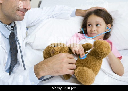 Ragazza distesa nel letto di ospedale, sorridente al medico come entrambi mantengono stetoscopi per la ragazza peluche Foto Stock