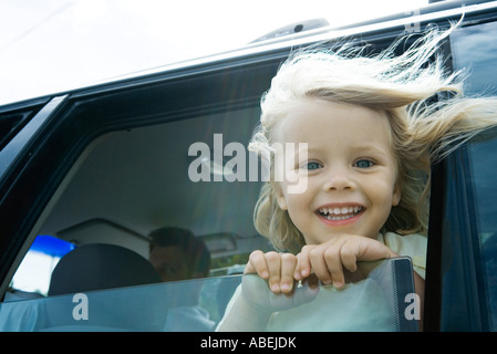 Bambina in auto, guardando fuori della finestra, sorridente in telecamera, capelli soffiando nel vento Foto Stock