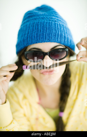 Giovane donna in maglia che indossa un cappello e occhiali da sole, tenendo premuto Fine della spiralina oltre il labbro superiore, ritratto Foto Stock