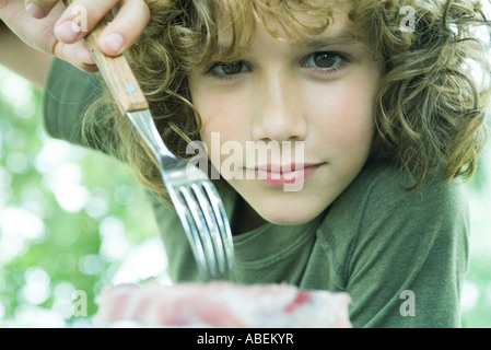 Ragazzo di prelevare il pezzo di carne con forcella, guardando la fotocamera Foto Stock