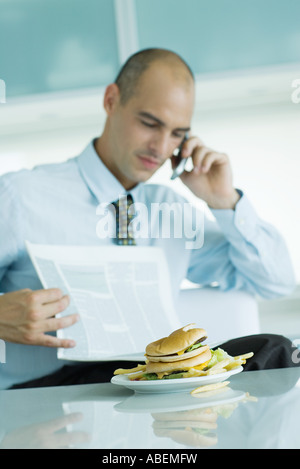 Uomo seduto a tavola con hamburger, leggendo il giornale e utilizzando il telefono cellulare Foto Stock