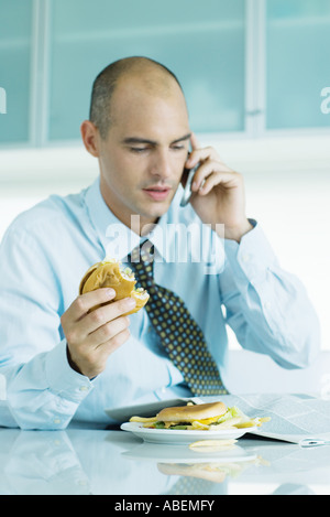 Uomo seduto a tavola a mangiare hamburger, leggendo il giornale e utilizzando il telefono cellulare