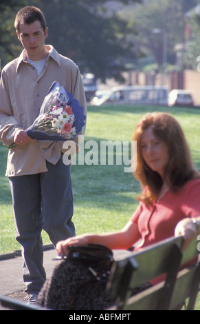 Apprensivi donna seduta su una panchina nel parco che si è avvicinato da giovane uomo che trasportano fiori Foto Stock