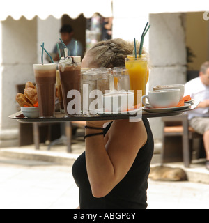 Cameriera che porta con una mano un grande vassoio di bevande pesanti verso i clienti al bar all'aperto, al tavolo del ristorante nella città vecchia di Corfù, isola greca di Corfù Foto Stock