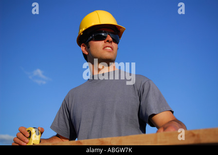 Operaio edile falegname in hard hat cercando durante la misurazione asse di legno con nastro di misurazione contro il cielo blu Foto Stock