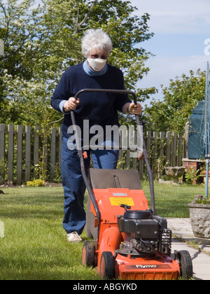 Donna anziana che falcia casa giardino prato domestico con un tosaerba mentre indossa una maschera di polvere viso per prevenire la febbre da fieno. Regno Unito Foto Stock
