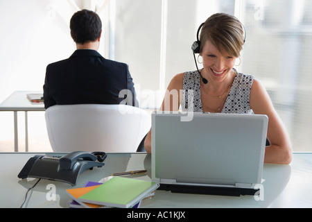 Business donna con cuffia lavorando sul computer portatile, collega di sesso maschile in background Foto Stock