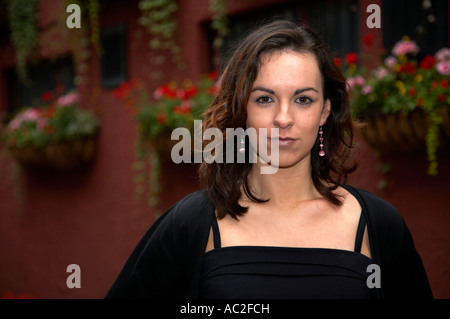 Testa e spalle di pretty irish brunette con occhi marroni e gli orecchini sorridente cercando di fotocamera con parete di cesti di fiori Foto Stock