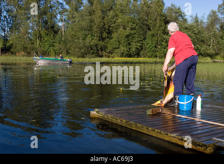 Donna anziana che lava tappeti su un molo in riva al fiume e un motoscafo / skiff che passa accanto , Finlandia Foto Stock