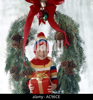 Brillantemente sorridente bambina tenendo un regalo di Natale è visto attraverso una grande ghirlanda di Natale in un inverno nevoso scena Foto Stock