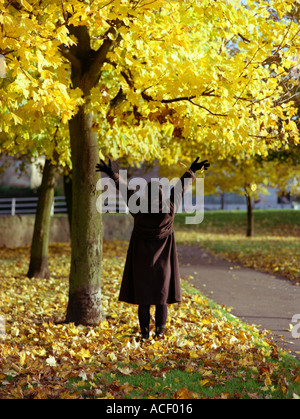 Giovane donna che indossa un guanto nero e un cappotto marrone che si allunga fino ad un albero con foglie gialle in autunno, dove il terreno è coperto di foglie Foto Stock