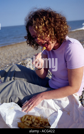 La donna a mangiare pesce e chip dalla carta con forcella di legno sulla spiaggia albrourogh Foto Stock