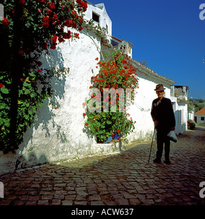 UOMO ANZIANO CHE CAMMINA CON UN BASTONE LUNGO UNA PARETE BIANCA CON FIORI ROSSI IN SALITA, ALTE VILLAGE, ALGARVE, PORTOGALLO, EUROPA Foto Stock