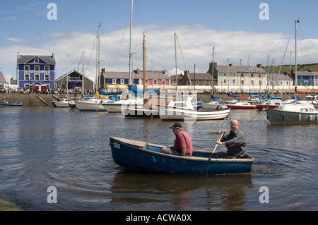 Due uomini in piccola barca a remi (OFFERTA) sull'acqua Aberaeron harbour estate mattina di sole con frutti di mare festival sul quay Foto Stock