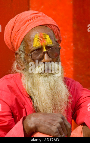 Chiudere verticale su ritratto di un anziano Sadhu nel colore rosso brillante abbigliamento e turbante seduto fuori un tempio Foto Stock