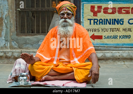 Ritratto orizzontale di un anziano Sadhu vestito in tradizionali abiti dello zafferano seduta con le gambe incrociate su un marciapiede. Foto Stock