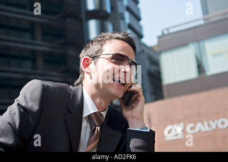Giovane imprenditore cercando di sorridere quando egli utilizza il telefono cellulare nella città di Londra seduti sui passi fuori Lloyds building Foto Stock