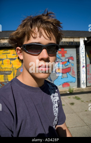 Ragazzo adolescente con disordinati capelli indossando occhiali da sole seduto fuori da un edificio coperto di graffiti colorati Foto Stock