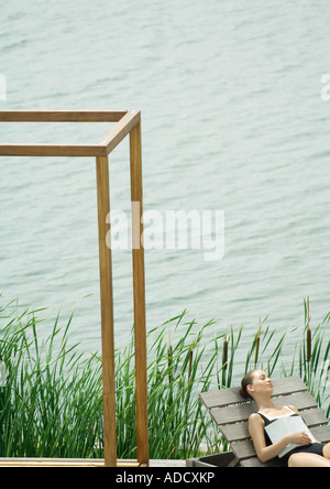 Donna sdraiata su poltrona accanto al lago, dormendo con libro sul petto Foto Stock