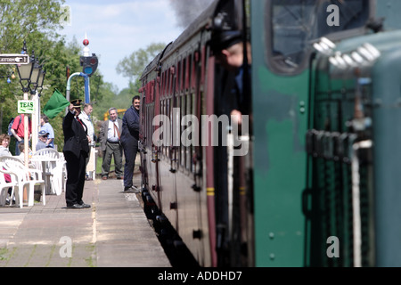 Treno a vapore circa per lasciare la stazione onde di guardia una bandiera verde e soffia il suo fischio Foto Stock