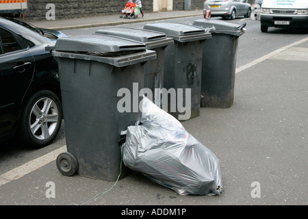 Sacchetti di plastica piena di spazzatura di overflow che giace accanto al grigio scomparti wheelie schierate sul sentiero dalla strada per la raccolta di rifiuti Foto Stock