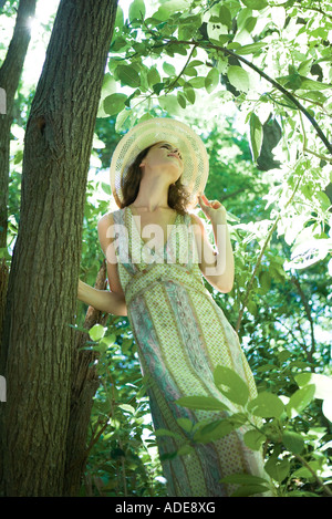 Giovane donna indossa sundress e cappello per il sole, in piedi accanto ad albero a basso angolo di visione Foto Stock